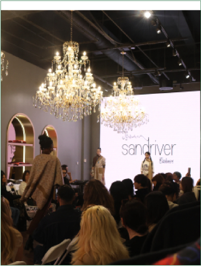 Runway presentation beneath chandeliers, with a seated audience and Sandriver backdrop.