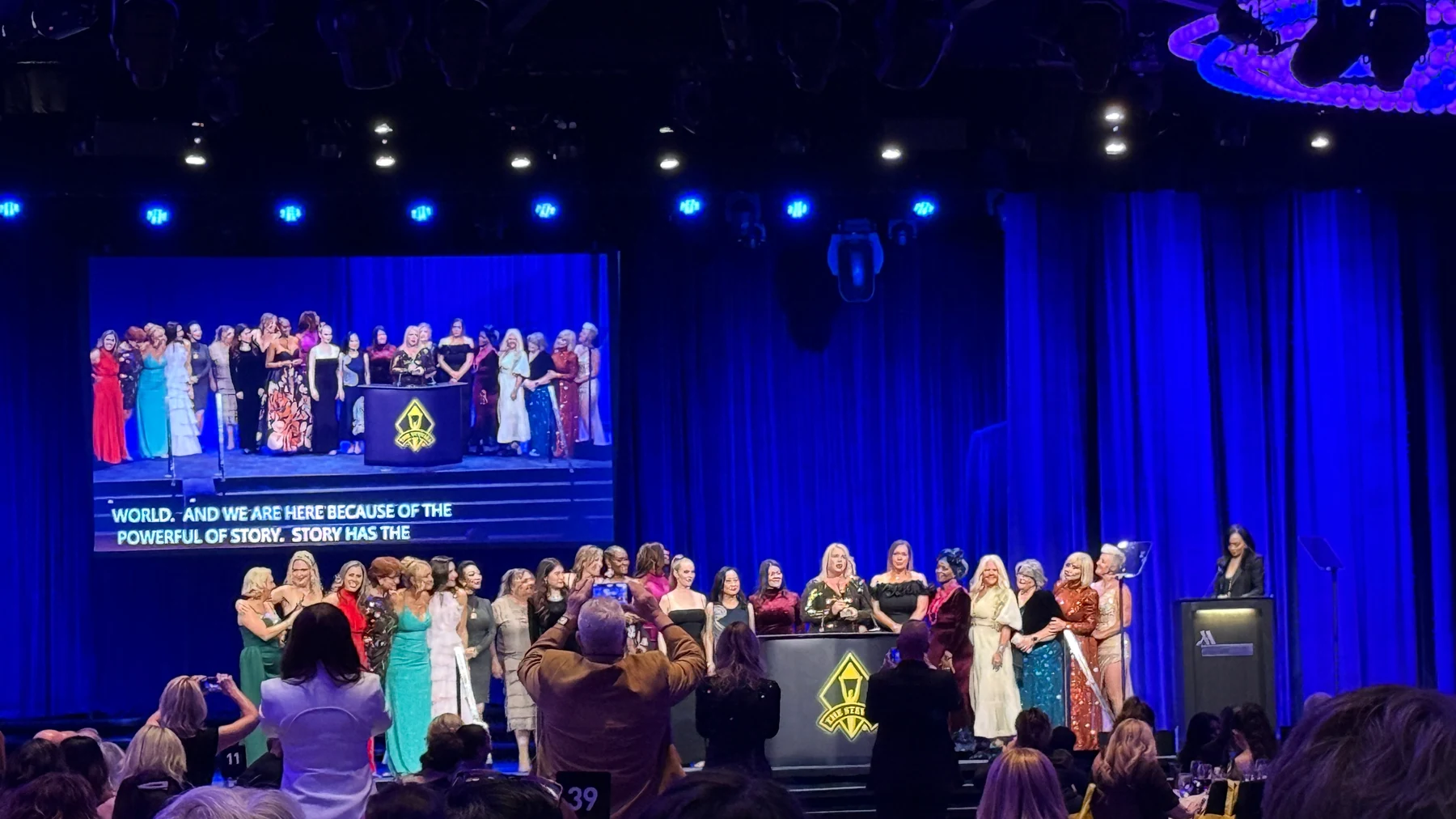 Stevie Awards ceremony with a group on the blue-lit stage and an audience in the foreground.