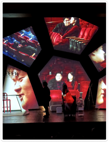 Central performers framed by polygonal projections of red cinema seats and faces on a dark theatre stage.
